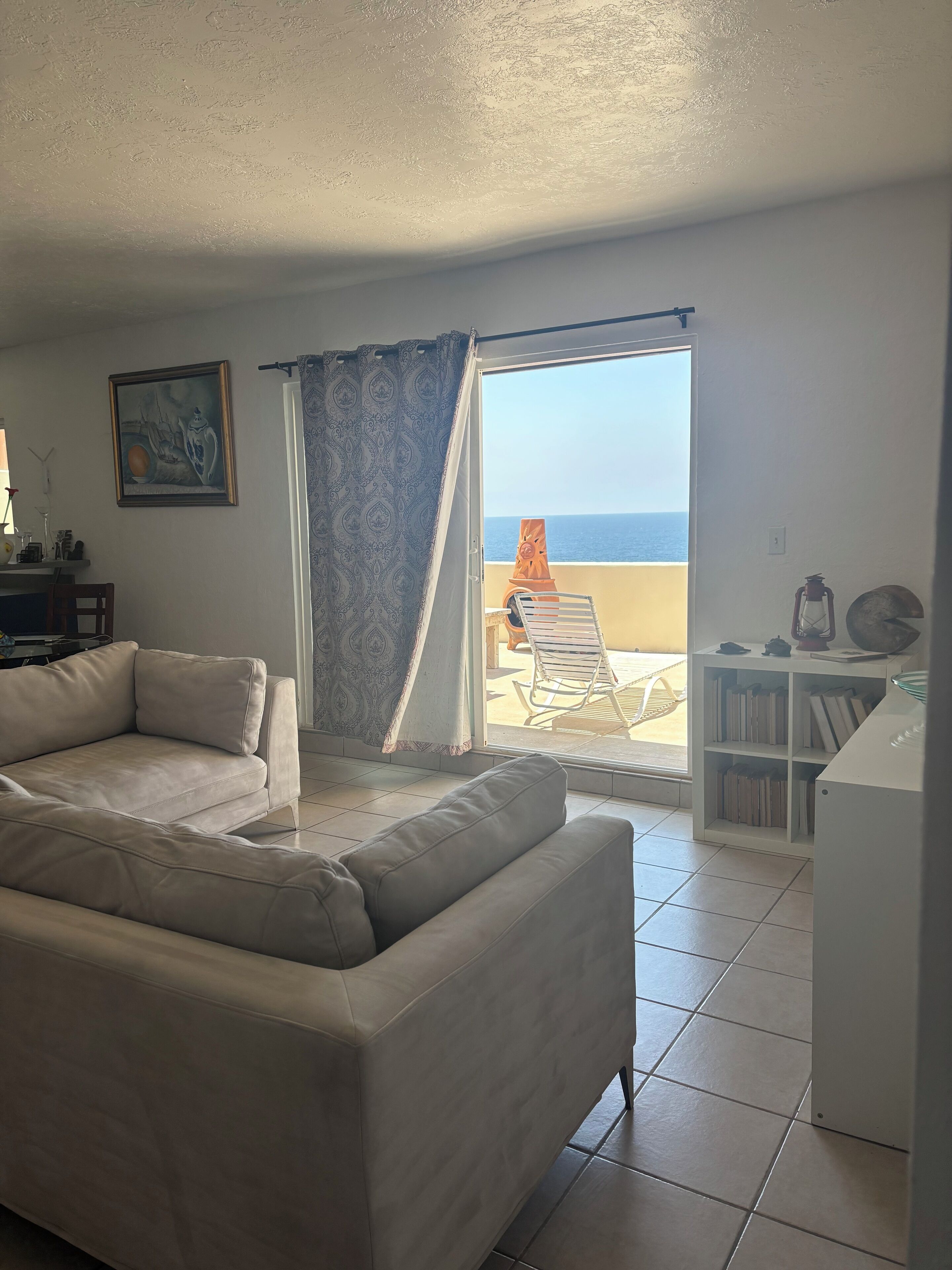 Una sala de estar con un sofá color crema, un cuadro y vista al mar a través de un ventanal corredero.
