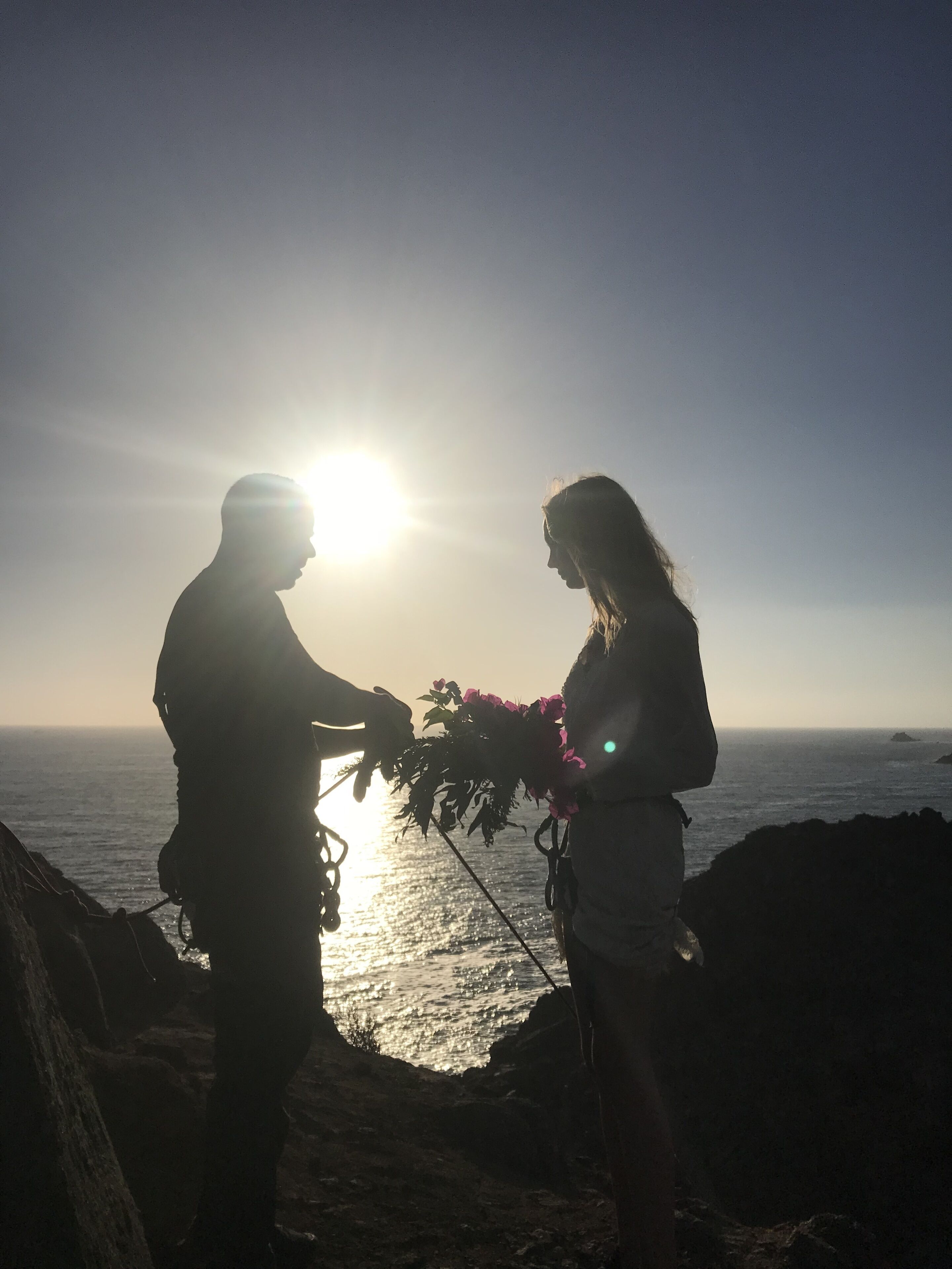 Twee silhouetten staan op een rotsachtige kustlijn tegen een ondergaande zon, met de oceaan op de achtergrond.