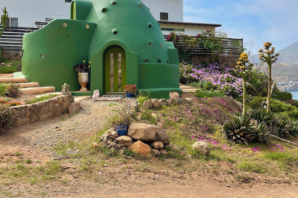 A green, dome-shaped building with a green door, surrounded by a garden with rocks and flowers.