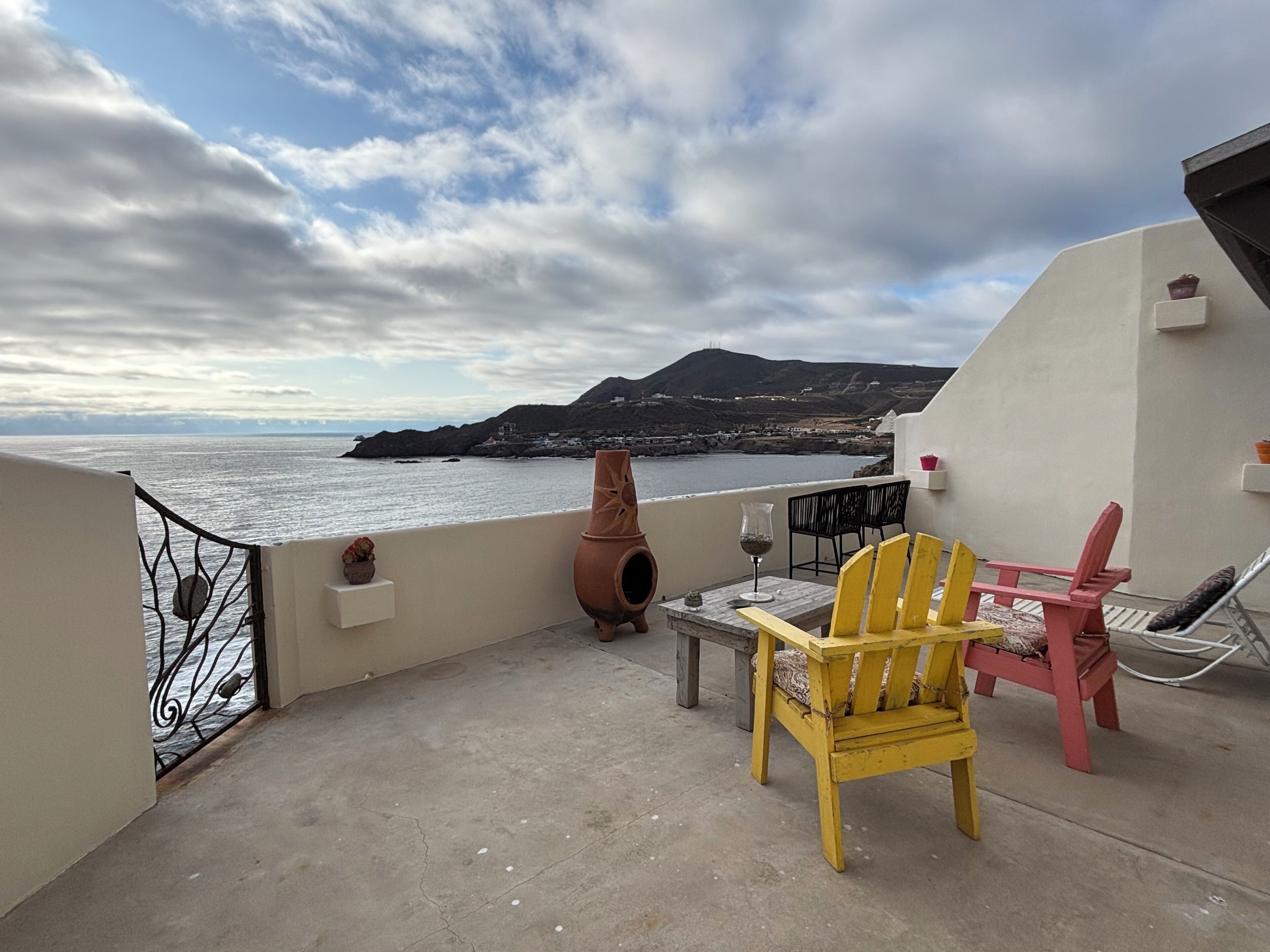 A balcony with a view of the sea and a mountain, featuring a table, chairs, and a potted plant.