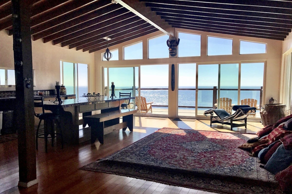 A spacious living area with a large window overlooking the ocean, a wooden floor, and a cozy seating area with a rug.