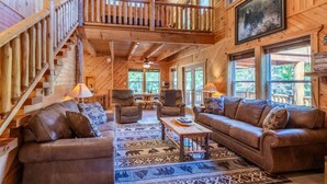 A wooden cabin interior with a central seating area, a staircase, and large windows. - Bearfoot's Nantahala Retreat-Pastoral View - Internet - Pool table - Fire Pit (Bryson City)