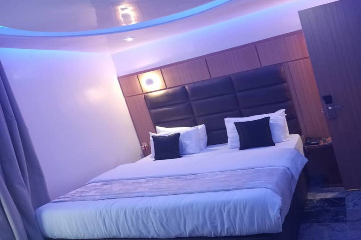 Quarto de hotel moderno com uma cama grande, cabeceira com iluminação embutida e uma pequena mesa de cabeceira.