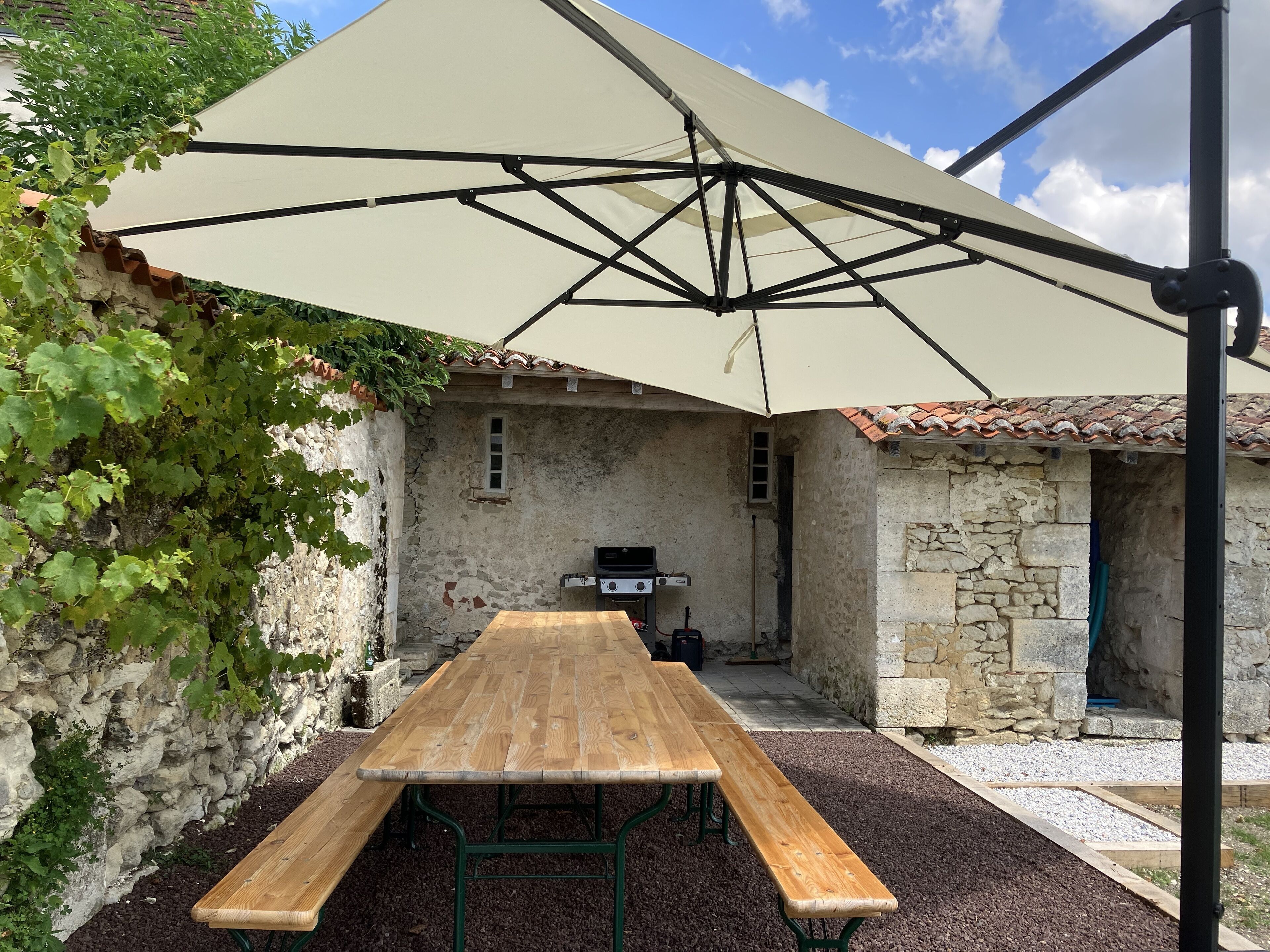 A rustic outdoor dining area with a wooden table and benches, a stone wall, and a pergola providing shade.