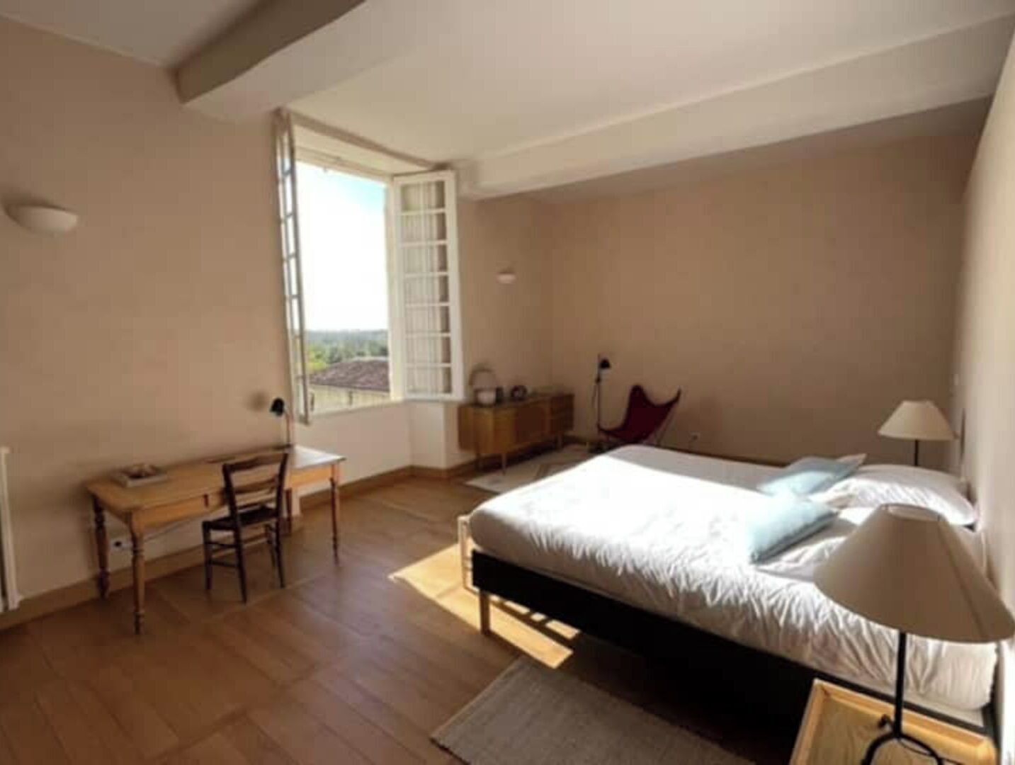 A bedroom with a bed, a desk, a chair, a nightstand, and a window with a view.