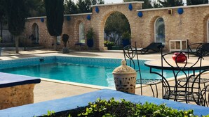 Piscina com parede de pedra e arcos, rodeada por assentos externos e plantas em vasos.