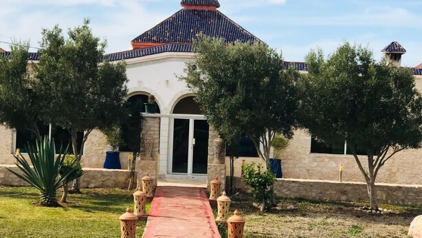 Um edifício com telhado em telhas, entrada central e um caminho que o leva até ele.