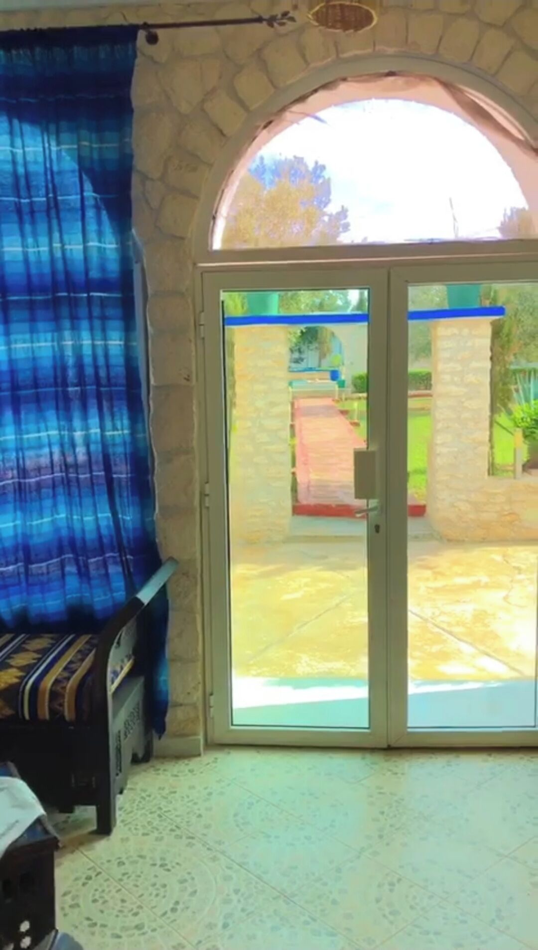 A room with a stone wall, a blue curtain, and a sliding glass door leading to an outdoor area with a pathway and greenery.