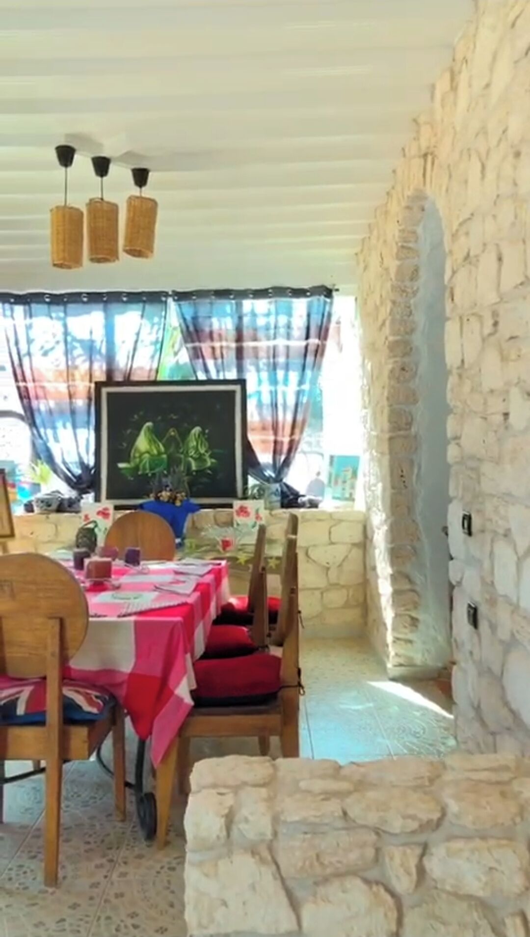 A dining area with a stone wall, a table with a red tablecloth, wooden chairs, and hanging lights.