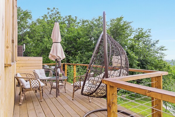 Una terraza de madera con mobiliario de mimbre, una silla colgante y una mesa con sombrilla.