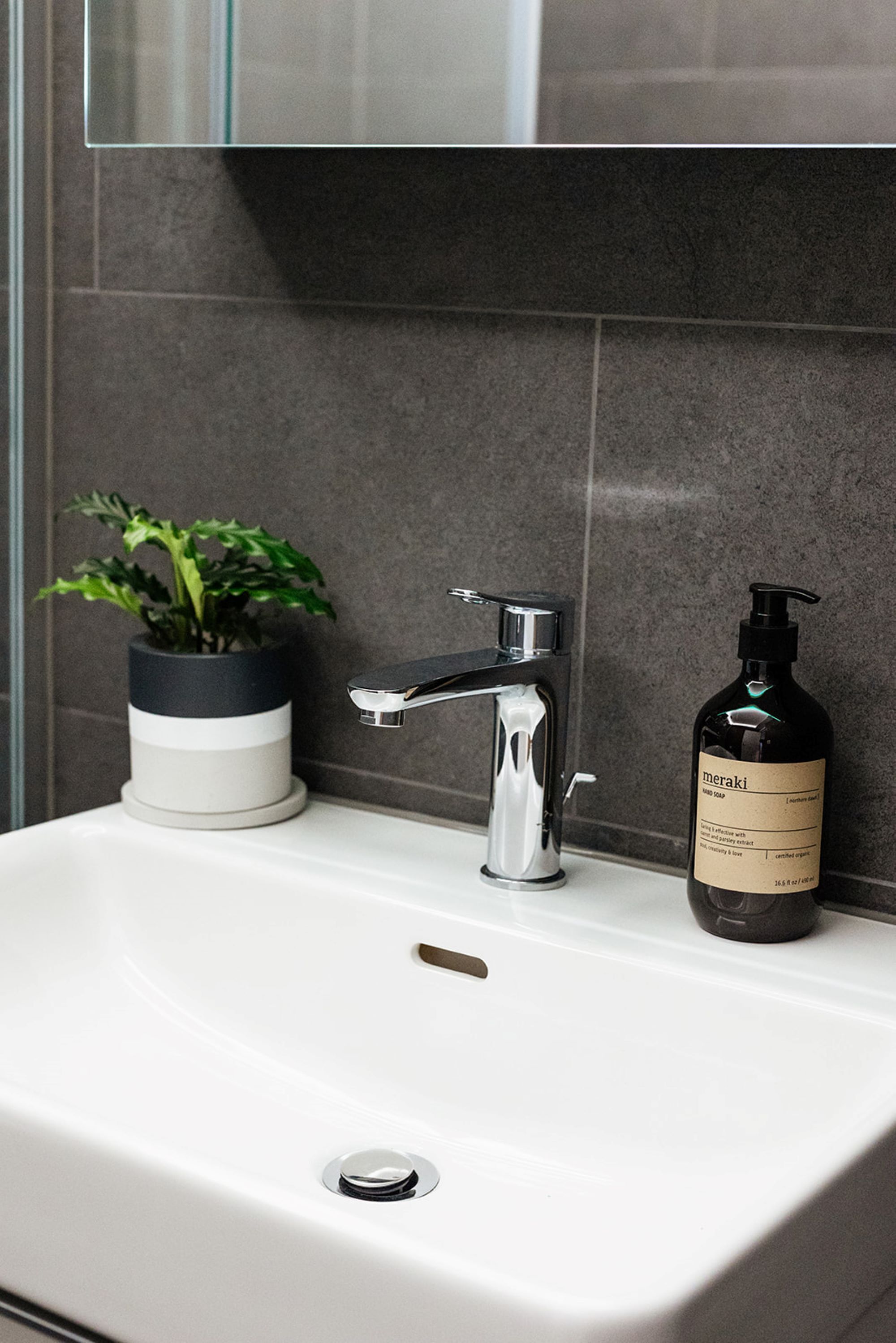 A bathroom sink with a modern faucet, a potted plant, and a bottle of Meraki hand soap.