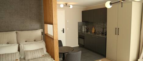 A compact bedroom with a bed, bedside tables, a small dining table, and a chair. Adjacent is a kitchenette with cabinets and a countertop.