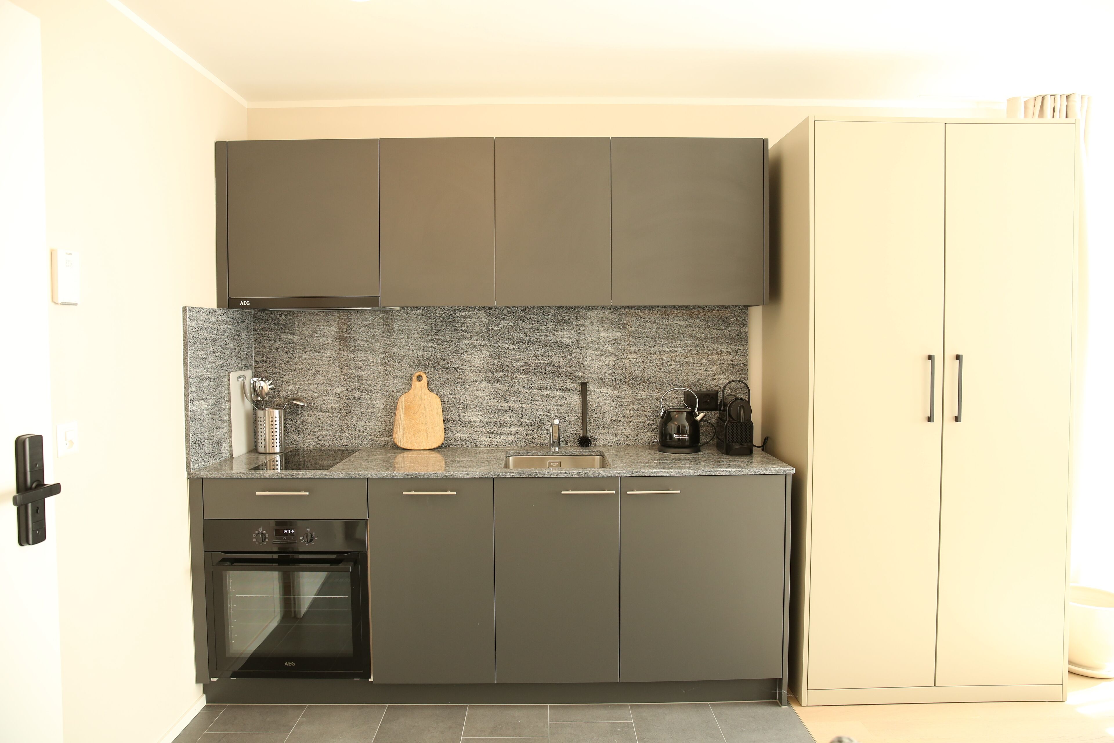 A modern kitchen with grey cabinets, a built-in oven, a granite countertop, and a beige wardrobe.
