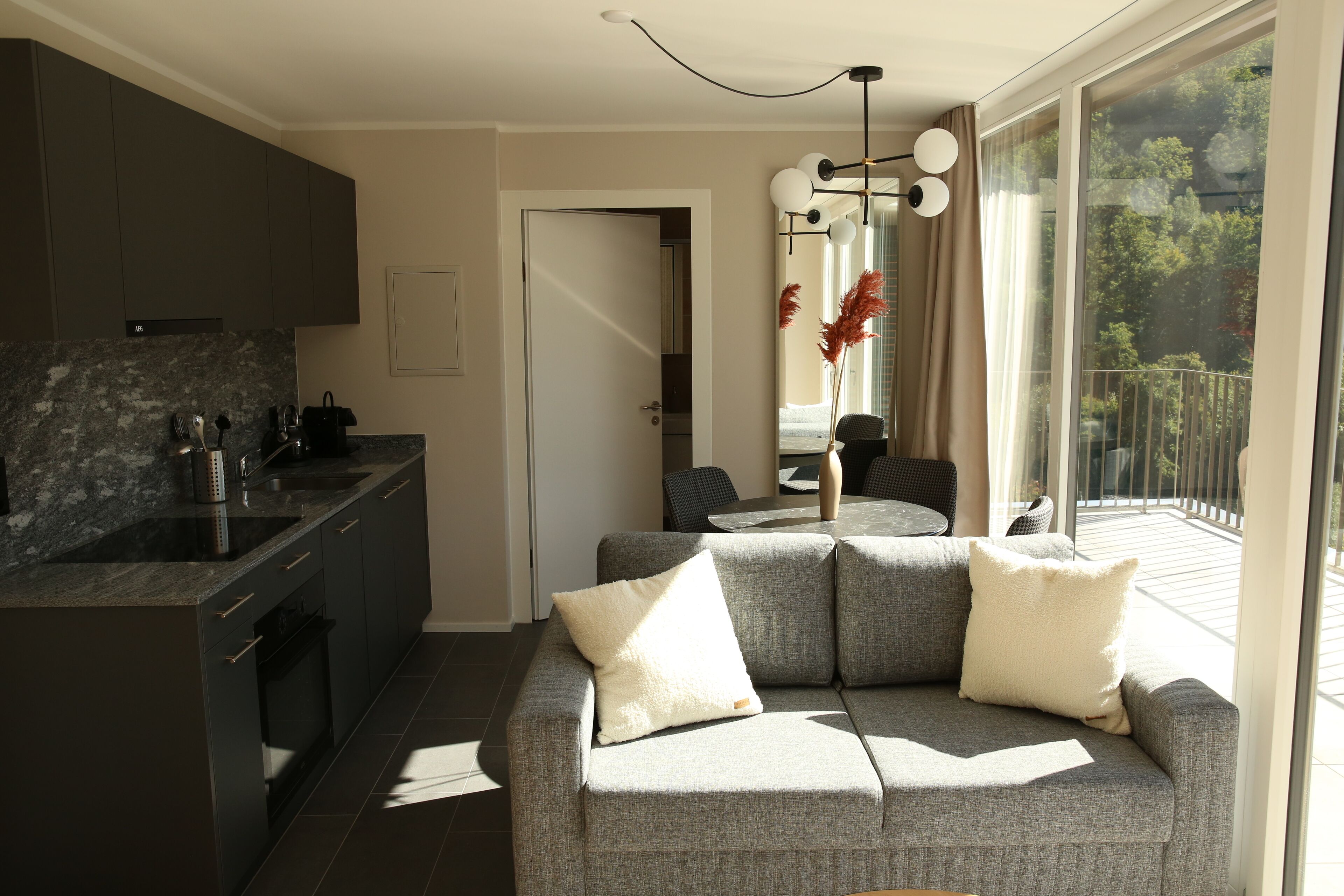 A modern kitchen with dark cabinets, a marble countertop, and a built-in oven. There is a grey sofa with white cushions in the living area, and a dining table with chairs. A large window provides a view of the outdoors.