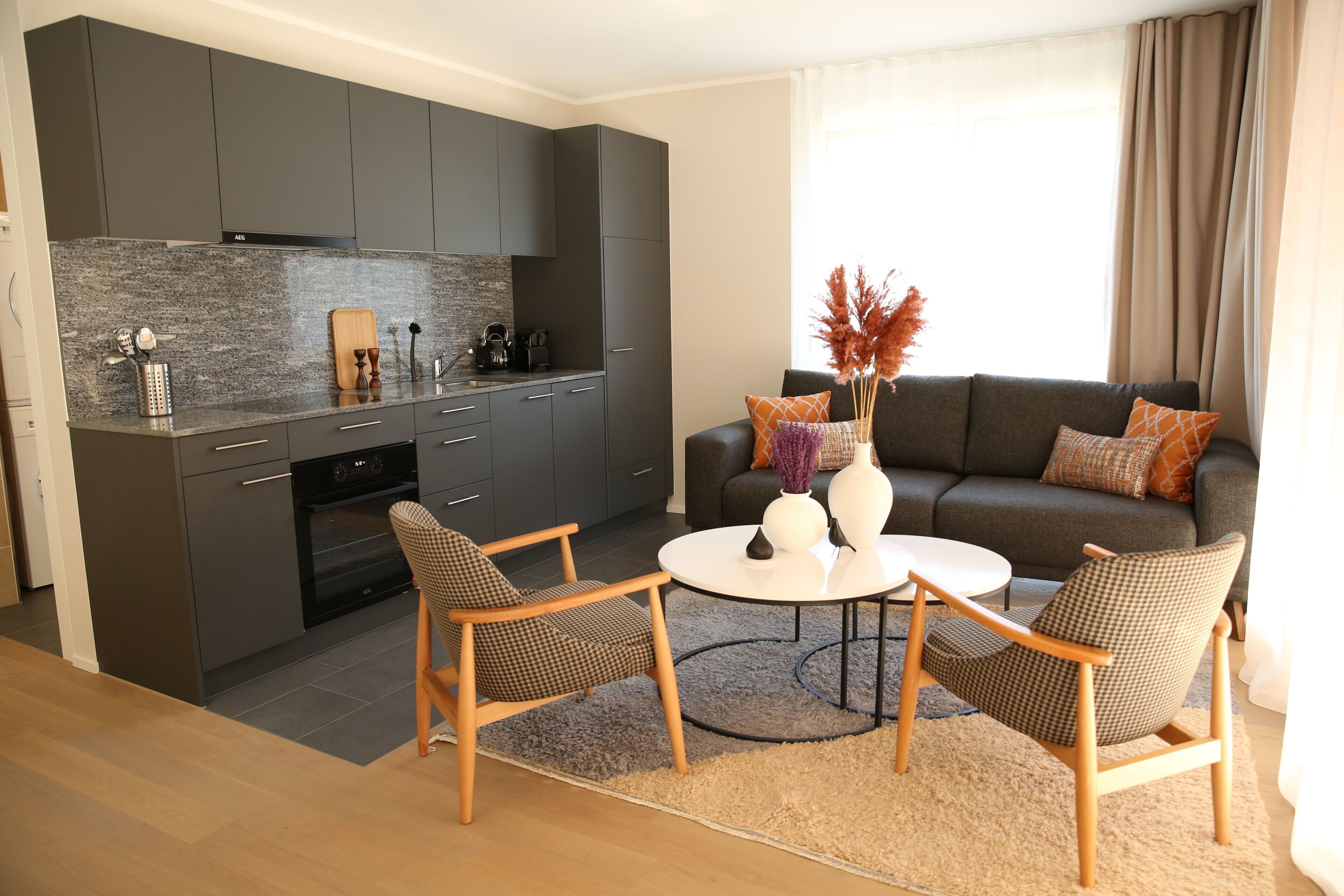 A modern kitchen with dark cabinets, a built-in oven, and a coffee machine. There is a dining area with a round table and two chairs. A sofa with cushions is placed near a window with curtains.