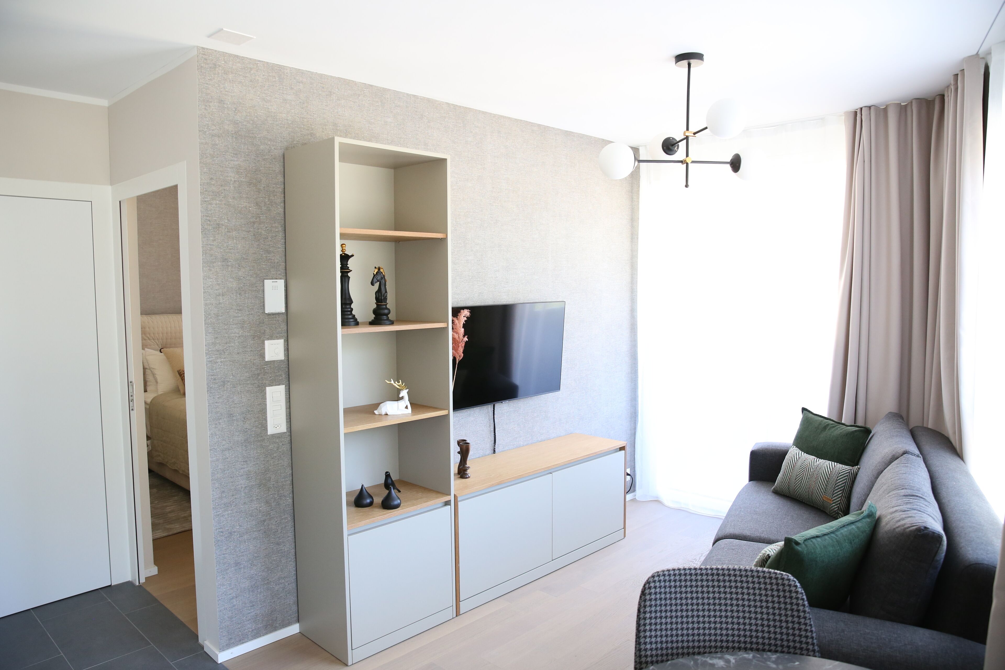 En moderne stue med en grå sofa, et fjernsyn monteret på væggen og et indbygget reolsystem med dekorative genstande.