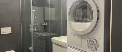 Bany compacte amb plat de dutxa envoltat de vidre, una rentadora Bosch i un vàter.