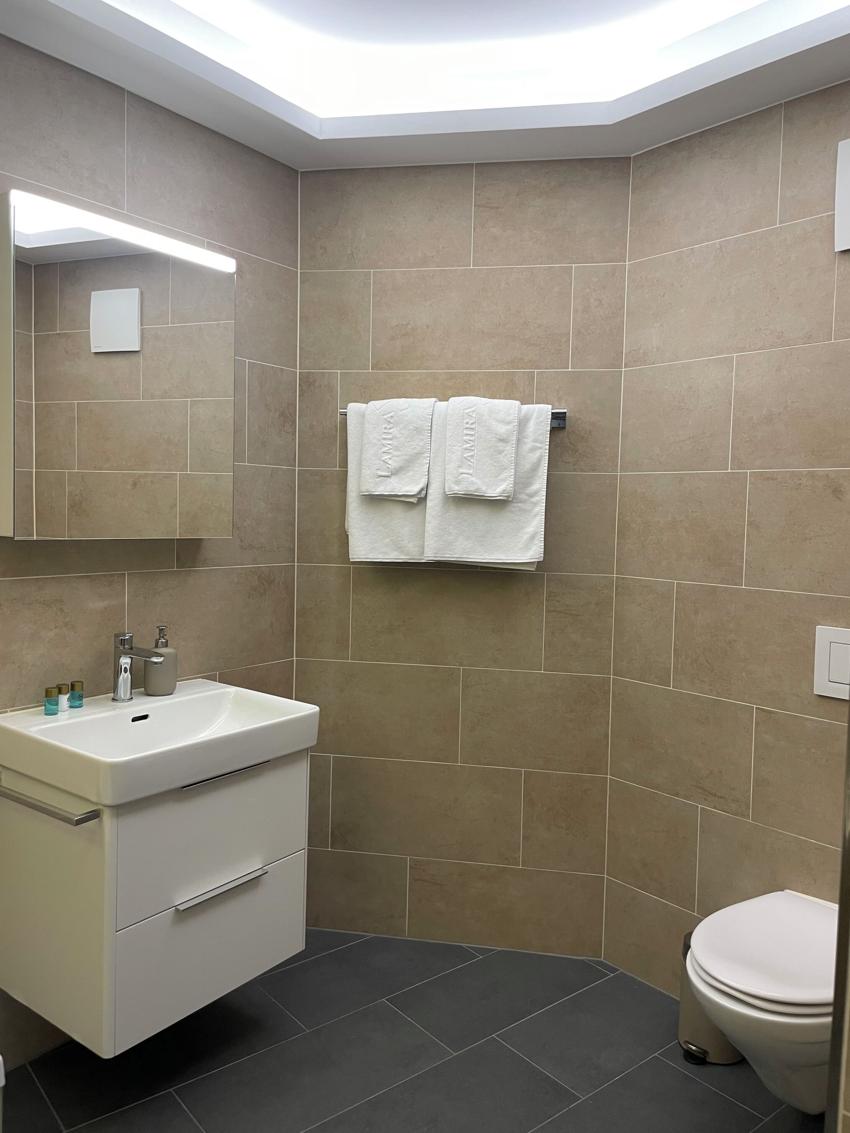 A modern bathroom with a white vanity, a mirror, a towel rack with two towels, and a toilet.