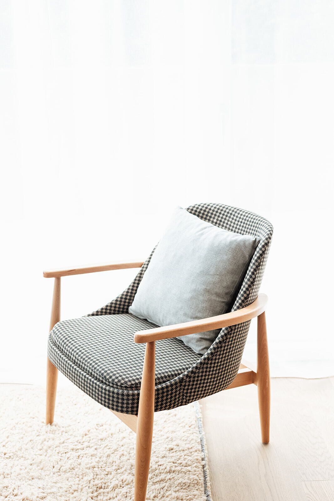 A houndstooth-patterned armchair with a light grey cushion, wooden armrests, and legs, positioned on a light-colored rug.