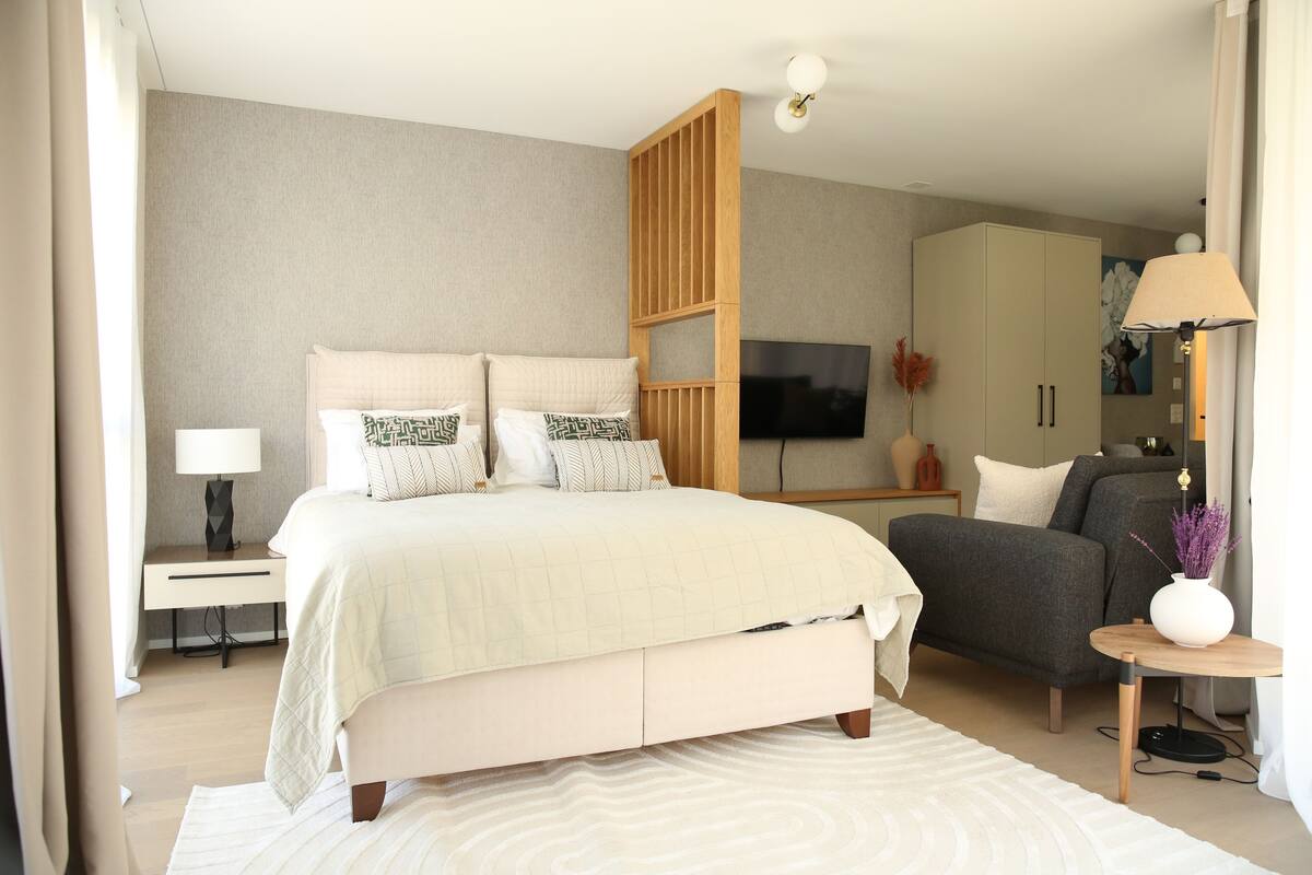 Une chambre moderne avec un grand lit, un canapé gris, une télévision et une cloison en bois.