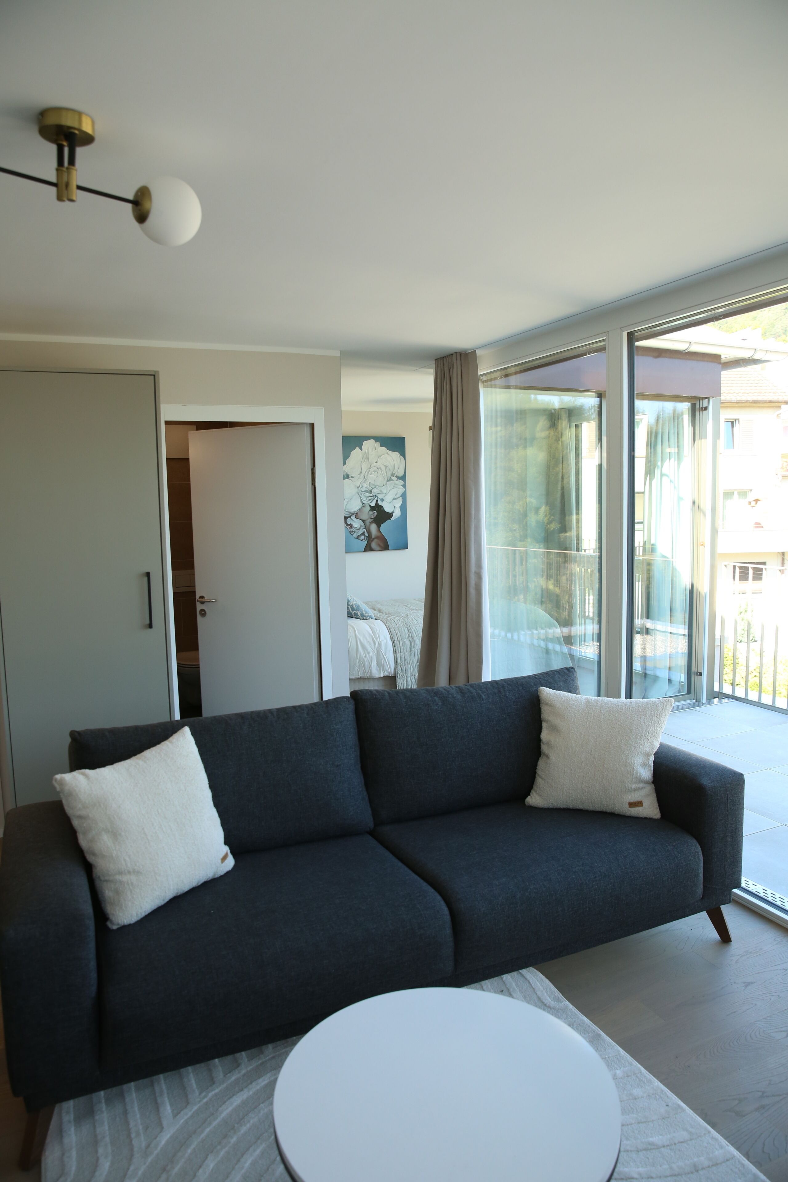 A modern living room with a dark grey sofa, a round white coffee table, and a large sliding glass door leading to an outdoor balcony.