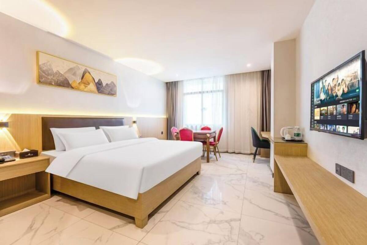 A hotel room with a bed, a desk, a chair, a table, a TV, and a painting on the wall.