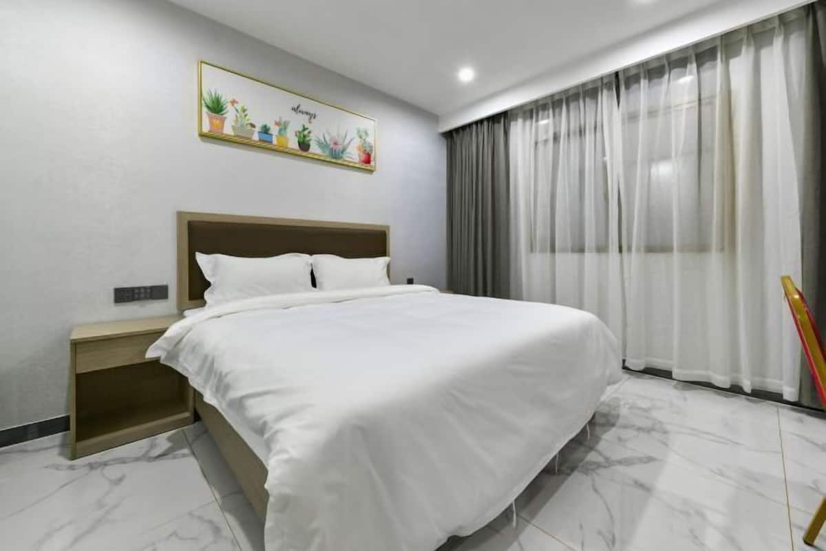 A modern hotel room with a large bed, a nightstand, a wall-mounted plant, and a window with curtains.