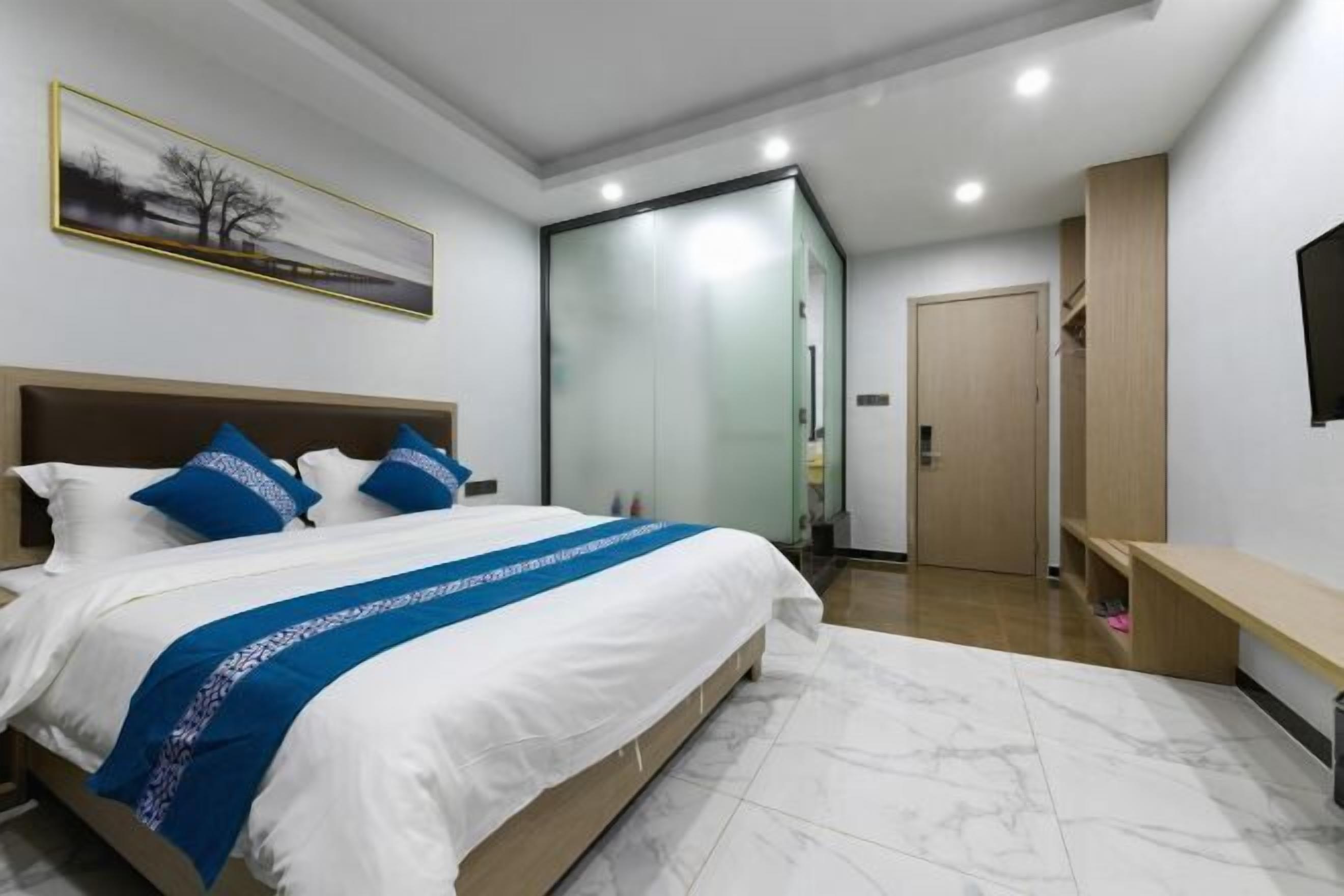 A modern hotel room with a large bed, a glass partition, a wooden door, and a bench.