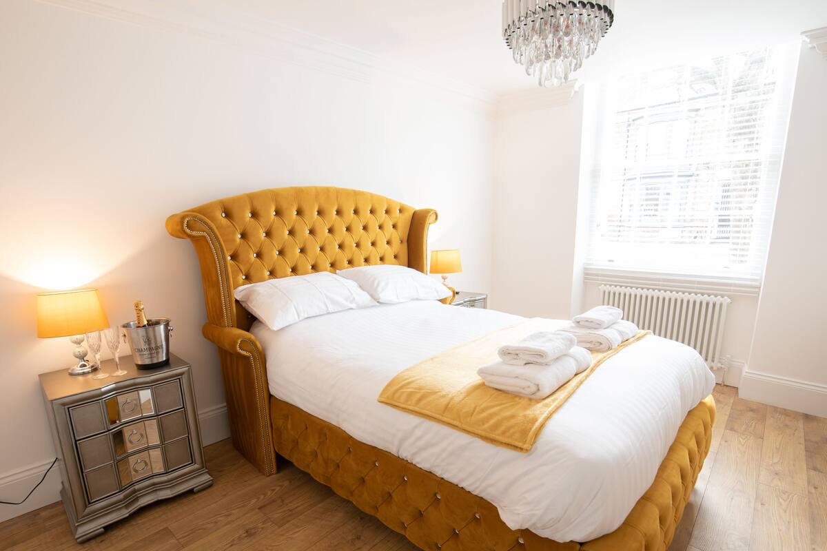 A hotel room with a large bed, a nightstand with a lamp, a minibar, and a window with blinds.