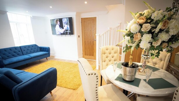A living room with a blue sofa, a yellow rug, a dining table with a champagne bottle, and a floral arrangement. - Ridley House Apartments (Yarm)