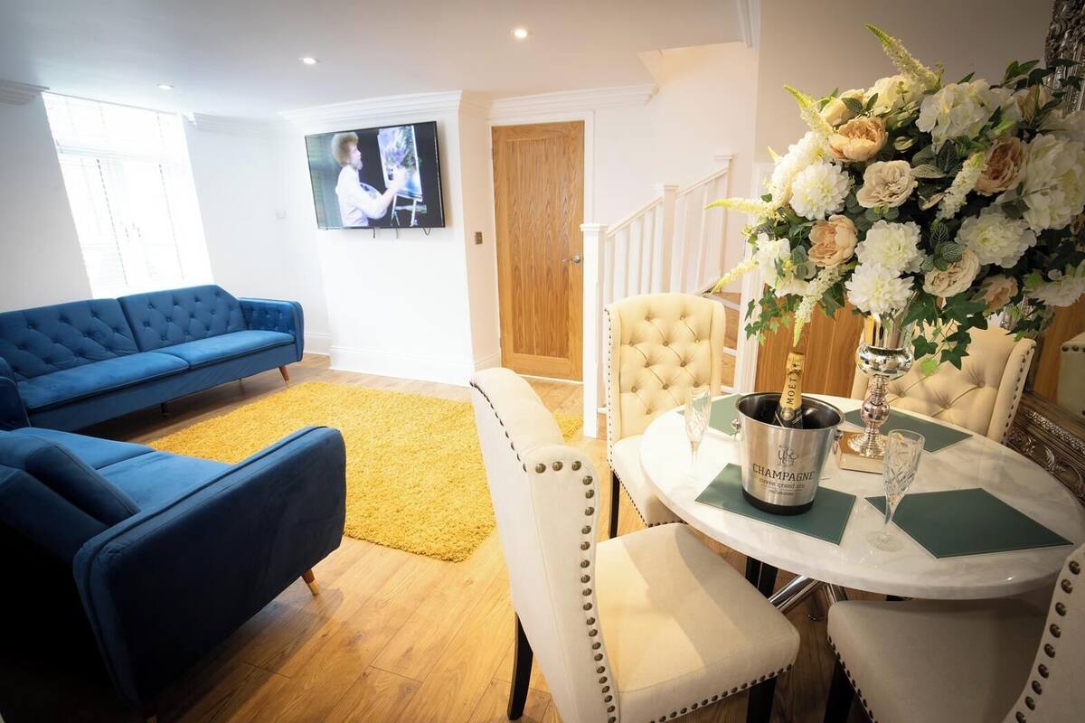 A living room with a blue sofa, a yellow rug, a dining table with a champagne bottle, and a floral arrangement.