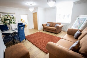 A living room with a brown sofa, blue armchair, and a red rug. There is a dining table with chairs and a vase of flowers. - Ridley House Apartments (Yarm)