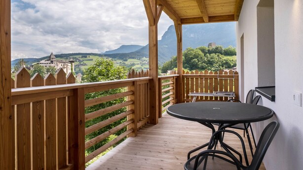 Een houten terras met een tafel en stoelen, met uitzicht op een schilderachtig landschap met bergen en een kasteel.