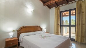 Une chambre avec une tête de lit en bois, une table de chevet avec une lampe, un lit recouvert d’une couvre-lit à motifs floraux et une vue à travers une porte vitrée.