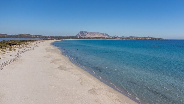 Une plage à l’eau claire, un mur de pierres et des montagnes à l’horizon.
