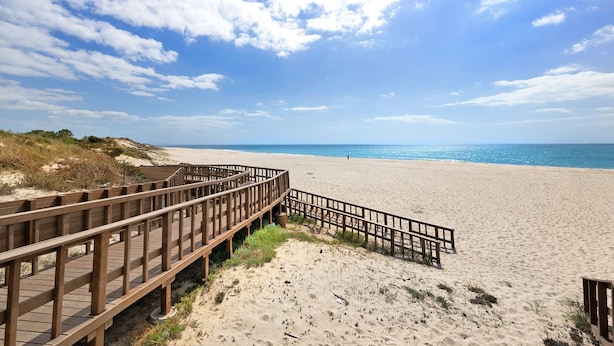 Ein hölzerner Steg führt zu einem Sandstrand bei blauem Himmel.