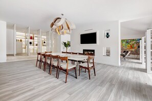 Un comedor moderno con una mesa y sillas de madera, una lámpara colgante original y un televisor grande en la pared.