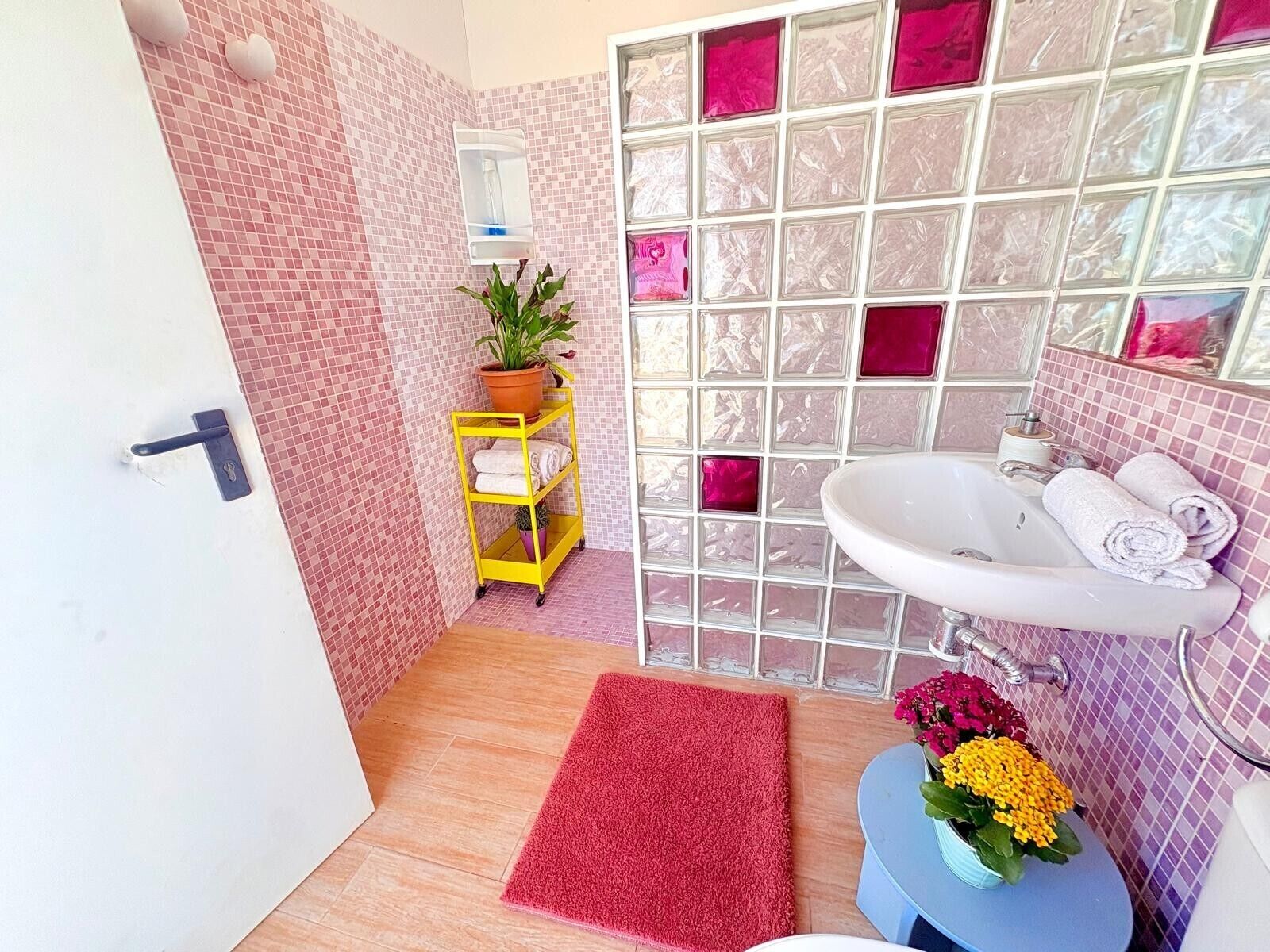 Un bagno con parete piastrellata, un lavandino, uno specchio, un carrello giallo, un tappeto rosso e una pianta in vaso.