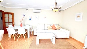 Uma sala de estar com sofá branco, mesa de jantar com cadeiras, lustre e ar condicionado instalado na parede.