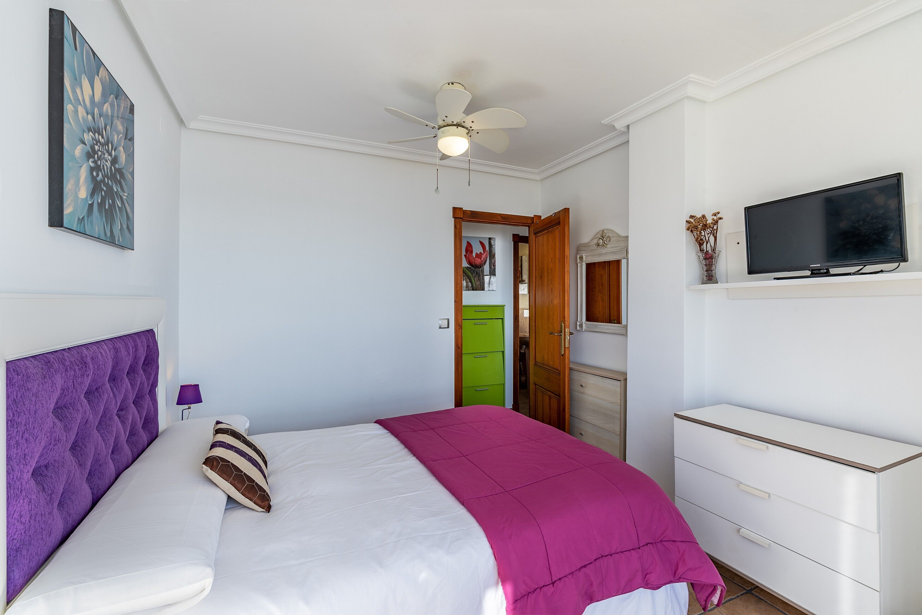 Een slaapkamer met een bed, een plafondventilator, een tv op een standaard en een deur naar een badkamer.