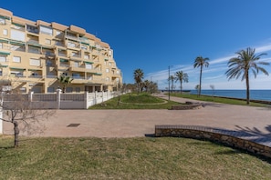 A multi-story hotel with balconies, a paved courtyard, palm trees, and a view of the sea.
