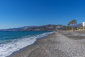 A pebble beach with a clear blue sky, a calm sea, and distant mountains.