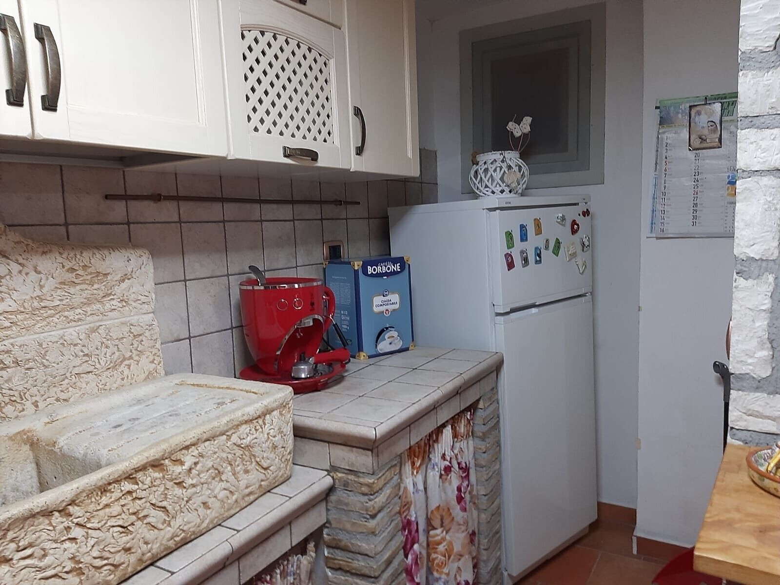 Una cocina con gabinetes blancos, una cafetera roja, un refrigerador con imanes y una ventana sobre el fregadero.