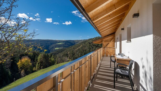 Een balkon met houten spijlen, een tafel met stoelen, en uitzicht op een bebost dal.