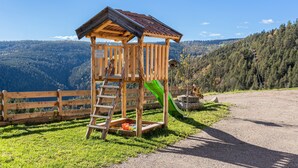 Een houten speelhuisje met een glijbaan, een ladder en een bordje, op de achtergrond van een berglandschap.