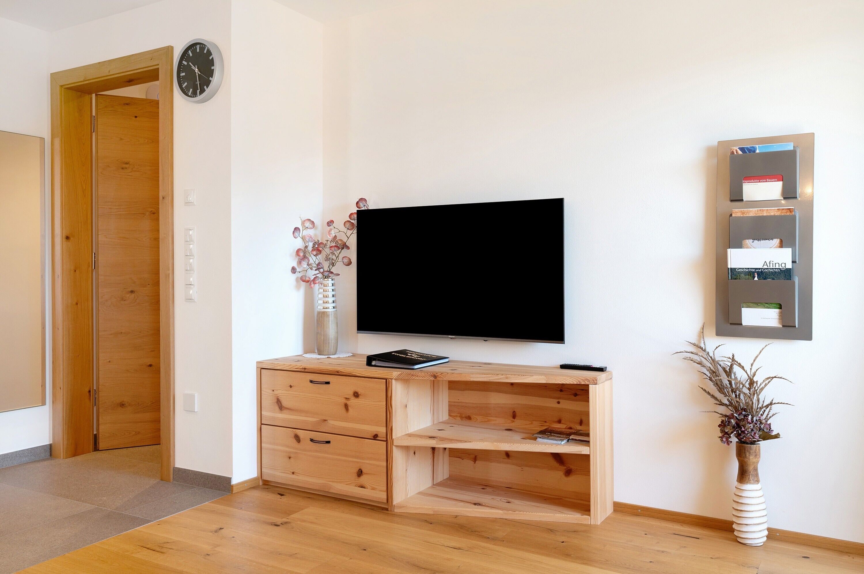 A room with a wooden desk, a flat-screen TV, a clock, a vase with dried flowers, and a framed photo on the wall.