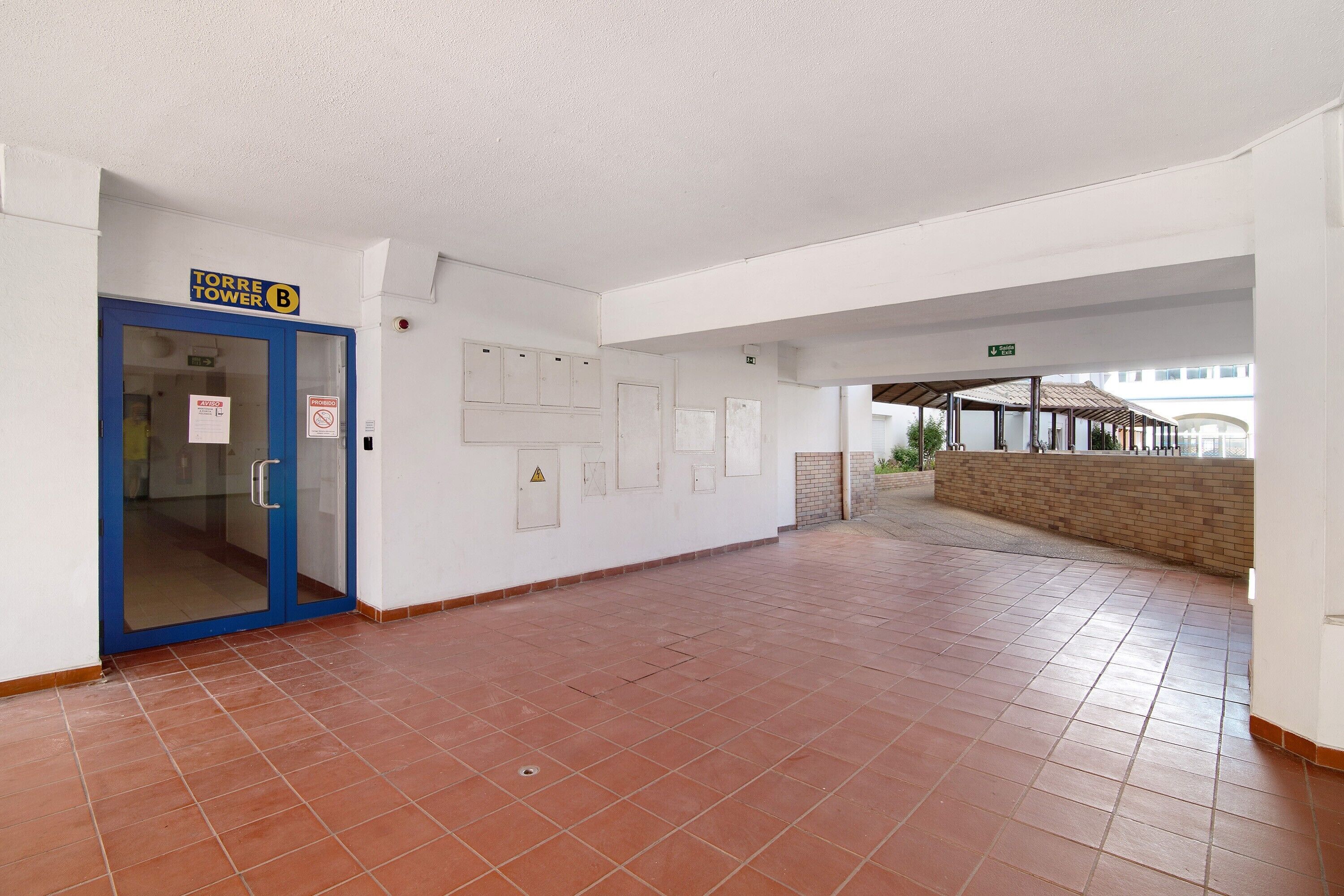Un pasillo con una puerta azul que dice 'TORRE TOWER B', suelo de baldosas y un área exterior cubierta.