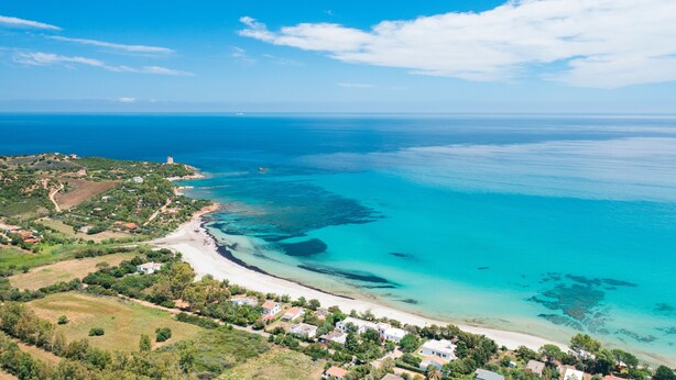 Une vue aérienne d’une zone côtière avec une plage, des eaux d’un bleu limpide et quelques maisons dispersées.