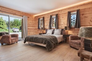 Quarto com painéis de madeira, uma cama, duas poltronas, uma mesa de cabeceira e uma janela com cortinas.