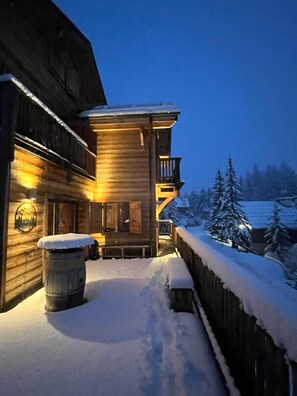 Una cabaña de madera con balcón, terraza cubierta de nieve y un barril de madera.